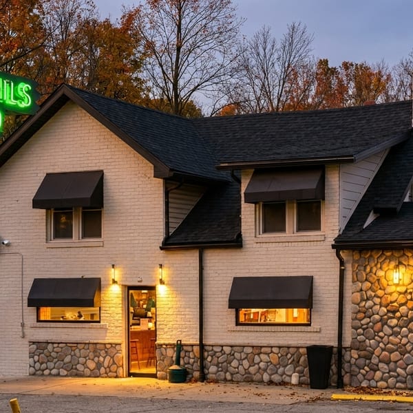 Exterior of a restaurant with a glowing green "COCKTAILS FOOD" sign, brick and stone facade, and autumn trees.