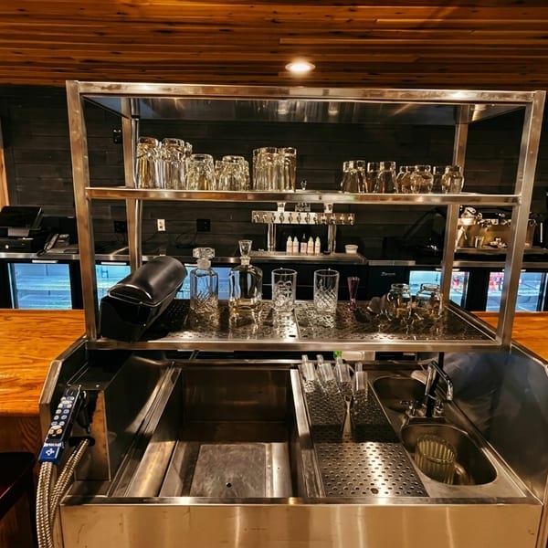 A well-lit, empty bar area with a wooden counter, stainless steel equipment, and glassware.