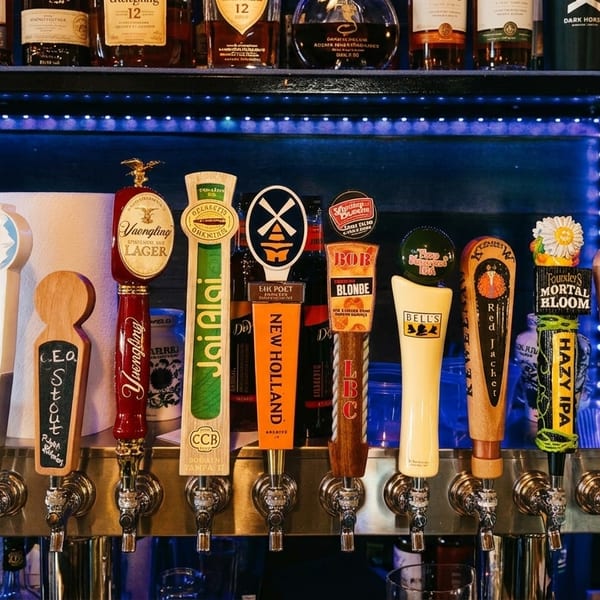 A row of diverse beer tap handles with various brewery logos, illuminated by blue lights.