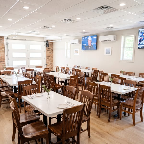 Bright, empty restaurant dining room with white tables, wooden chairs, and a large garage door window.